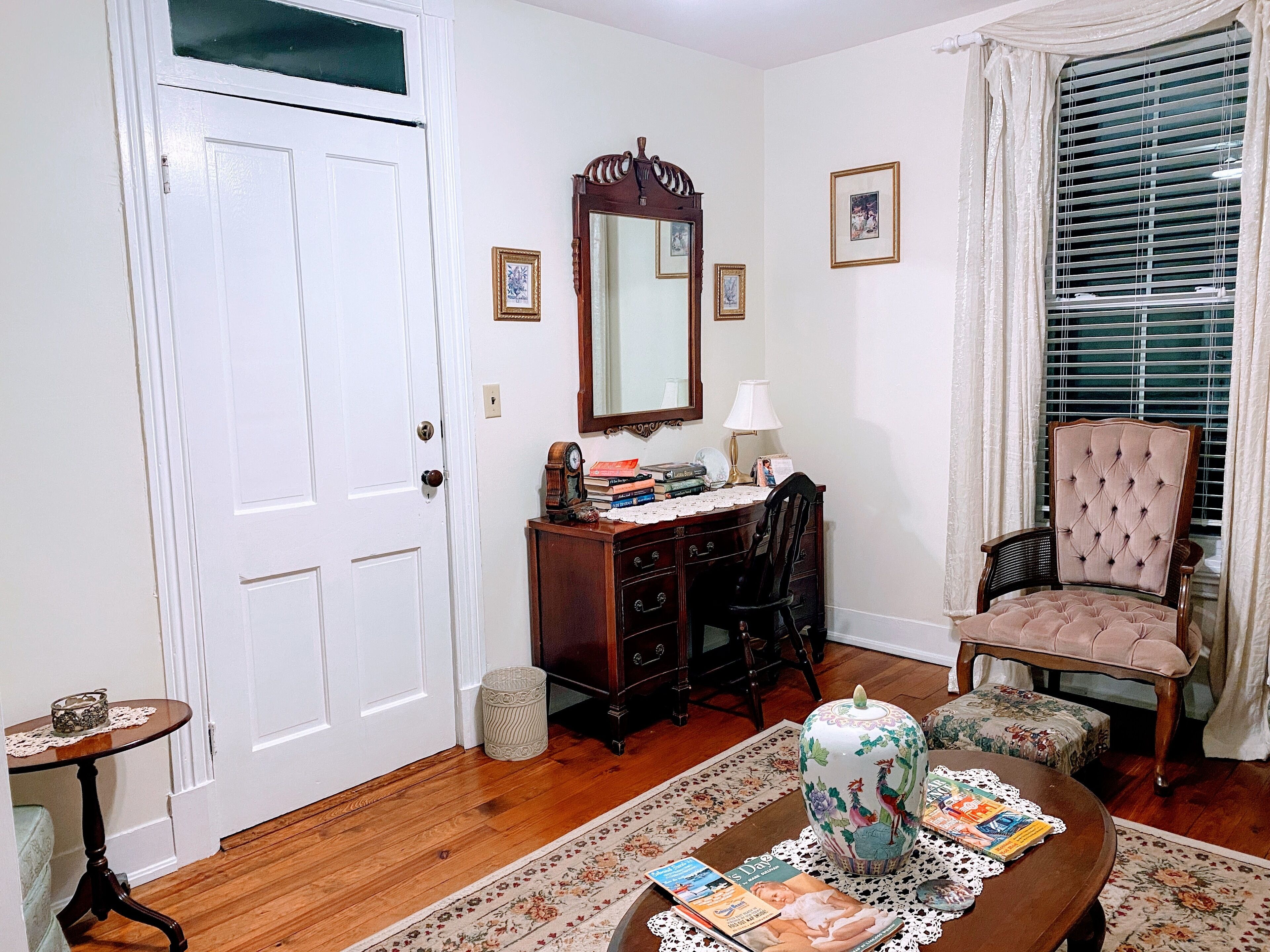 Una habitación con escritorio de madera, una silla, un espejo grande y un ventanal con persianas.