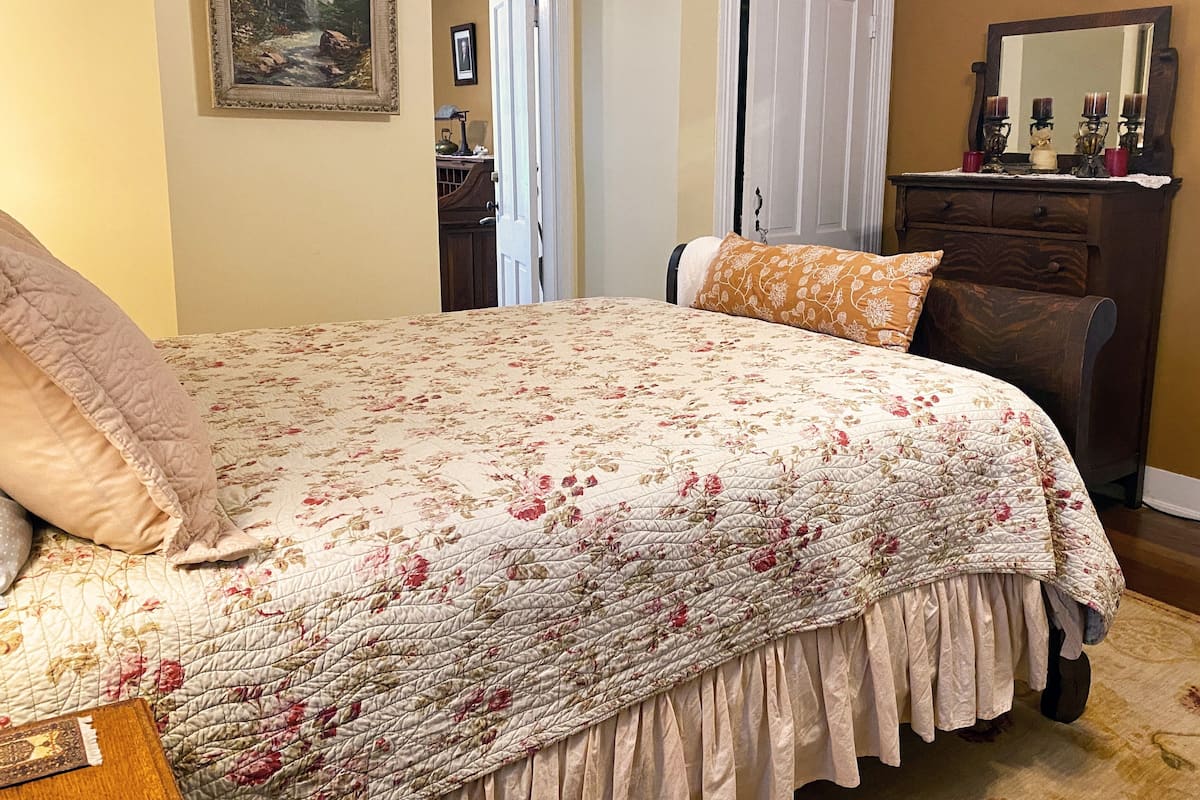 A bedroom with a floral bedspread, a wooden dresser, a mirror, and a door leading to another room.