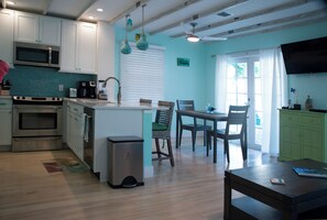 A kitchen with white cabinets, a microwave, an oven, and a coffee maker. There is a dining table with chairs and a trash can.