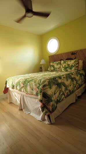 A bedroom with a bed, a ceiling fan, a round window, and a nightstand with a lamp.