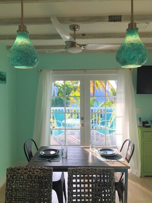 A dining area with a table set for two, two chairs, a ceiling fan, and a view through a glass door to an outdoor patio with chairs and a table.
