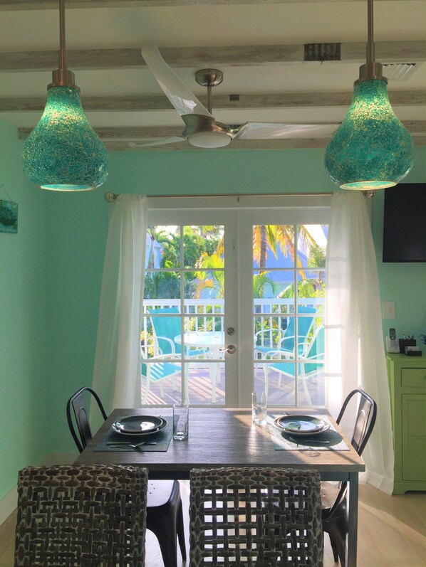 A dining area with a table set for two, two chairs, a ceiling fan, and a view through a glass door to an outdoor patio with chairs and a table.