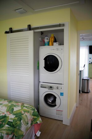 A laundry room with two stacked washing machines, a bed with a floral bedspread, and a kitchen area visible through an open door.