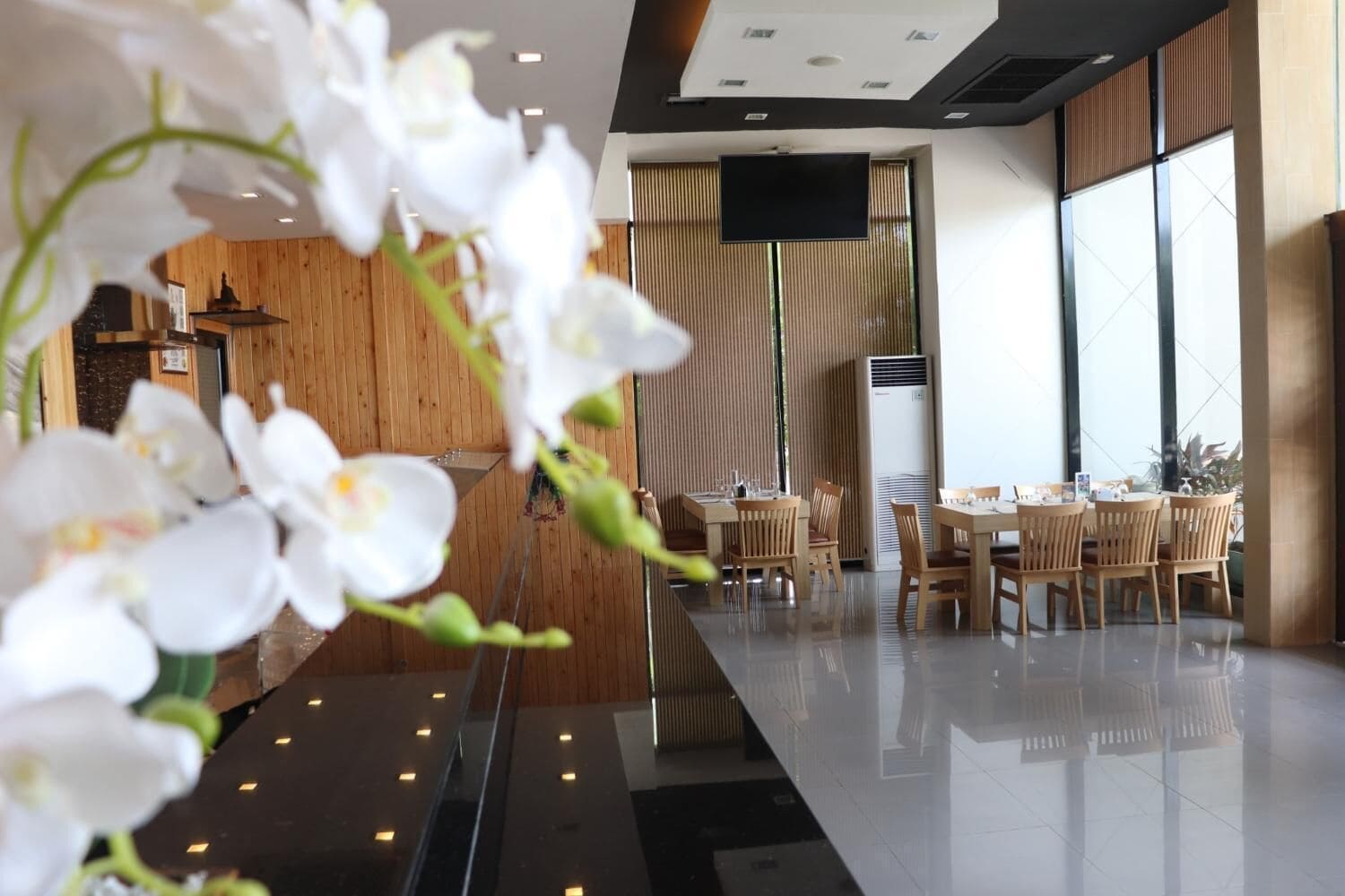 A modern hotel lobby with wooden paneling, a flat-screen TV, and a dining area with wooden tables and chairs.