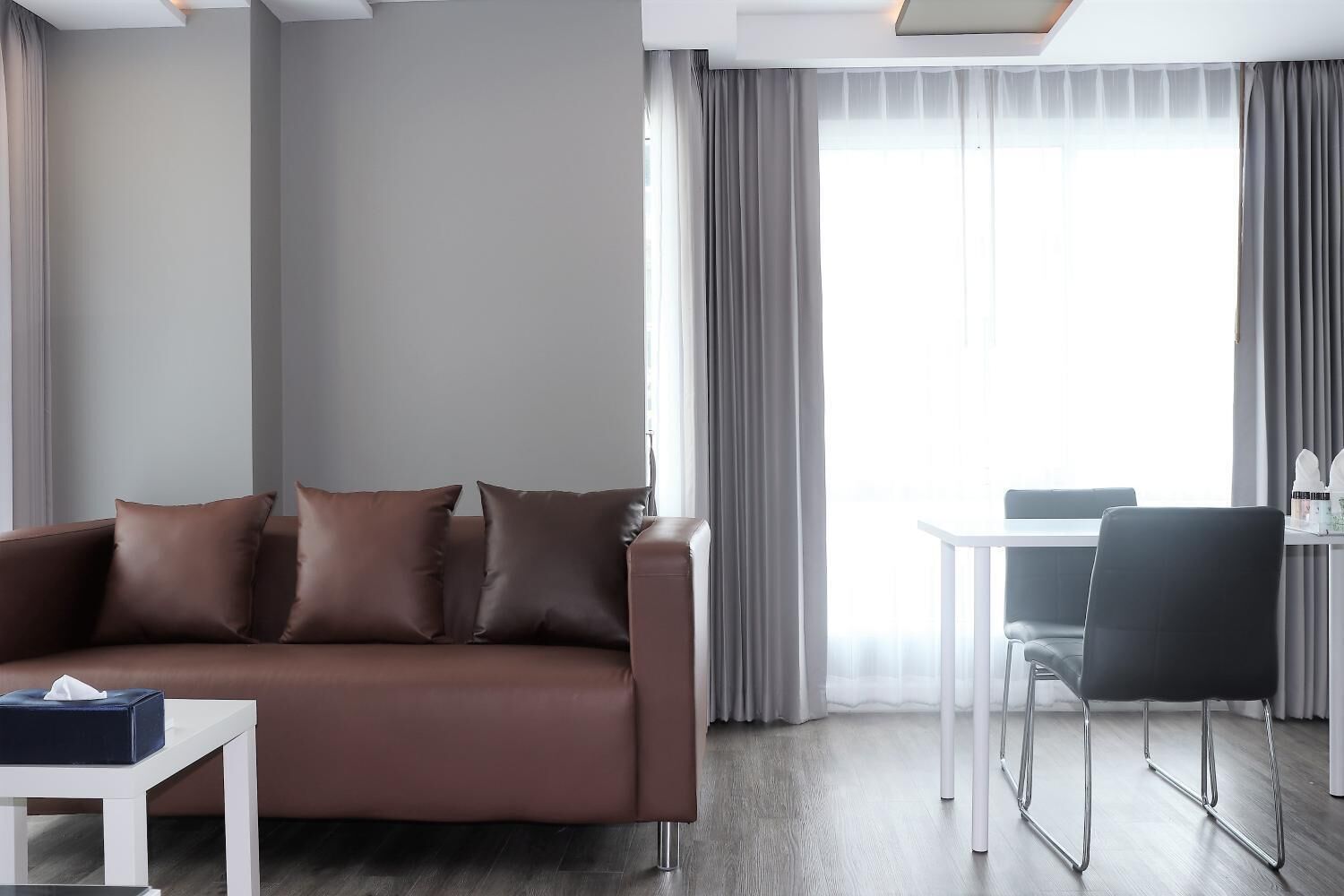 A modern living room with a brown sofa, a small table with a tissue box, a dining area with a white table and black chairs, and large windows with sheer curtains.