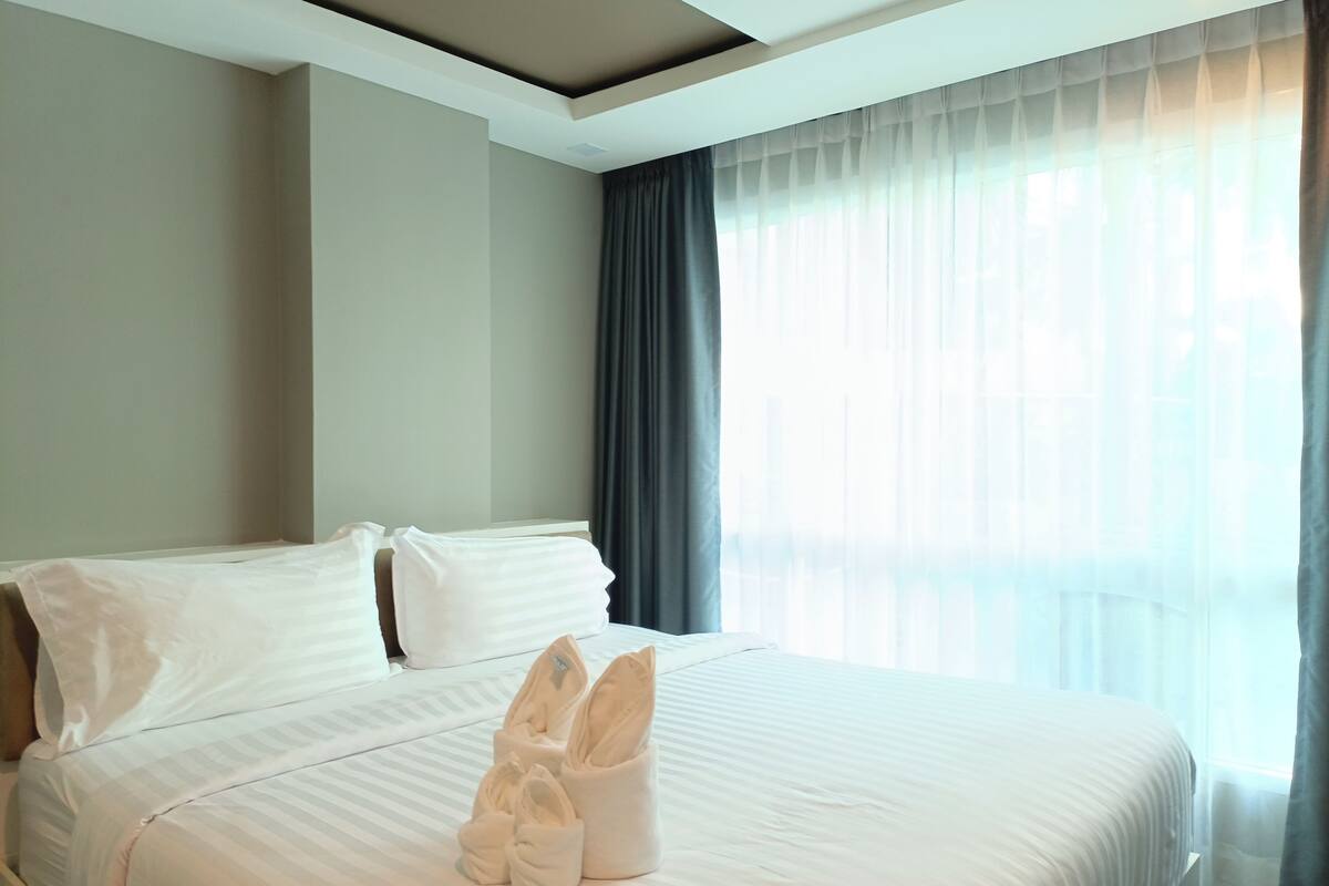 A hotel room with a neatly made bed, a window with sheer curtains, and a ceiling light.