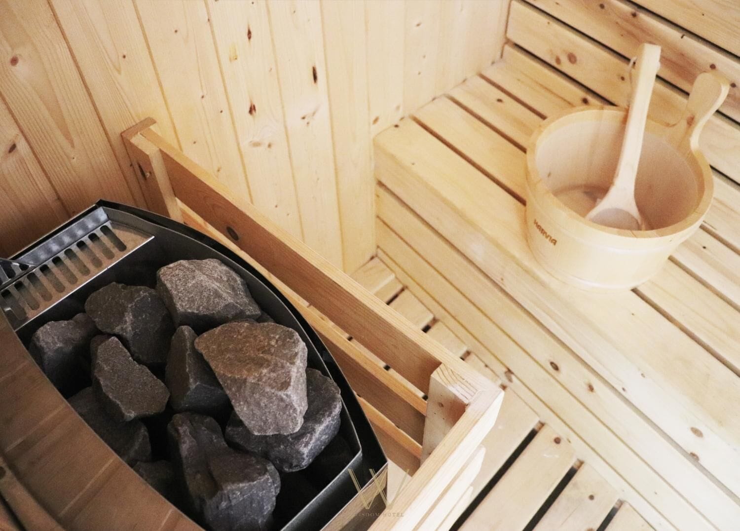 A sauna with wooden benches, a black container with rocks, and a wooden bowl with a ladle.