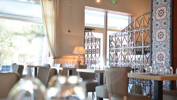 A restaurant with tables set for dining, a patterned wall, and a window with a view of greenery. - Hôtel Le Grillon (L'Île-Rousse)