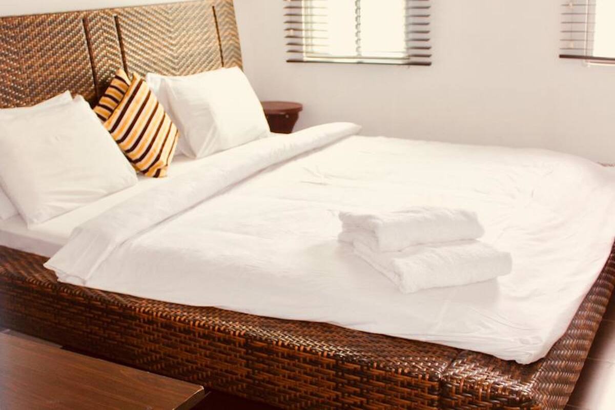 A neatly made bed with white linens and striped pillows, a woven headboard, and a wooden nightstand.