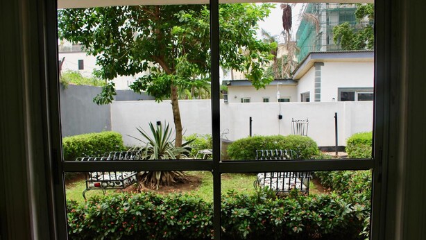 Une vue à travers une fenêtre donnant sur une cour intérieure avec une zone pavée, un arbre et un banc en métal.