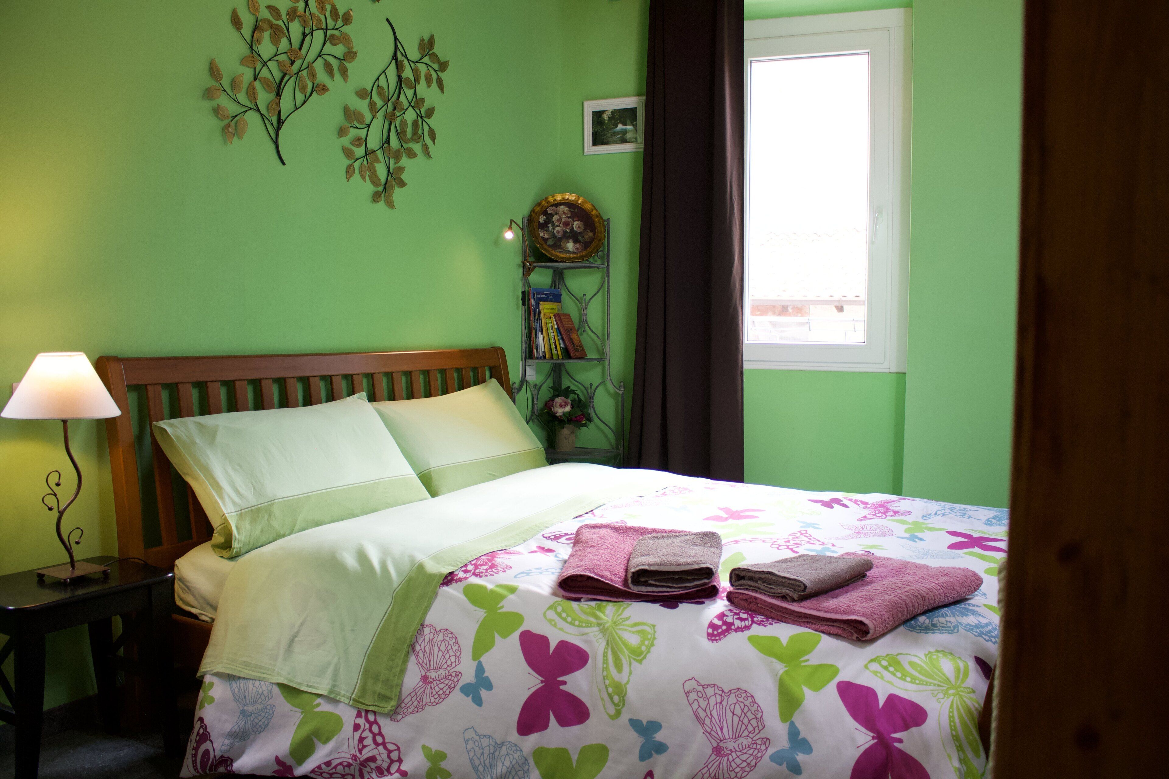 Una cama bien hecha con una colcha estampada, una mesita de noche con una lámpara y una estantería de pared con libros y objetos decorativos.