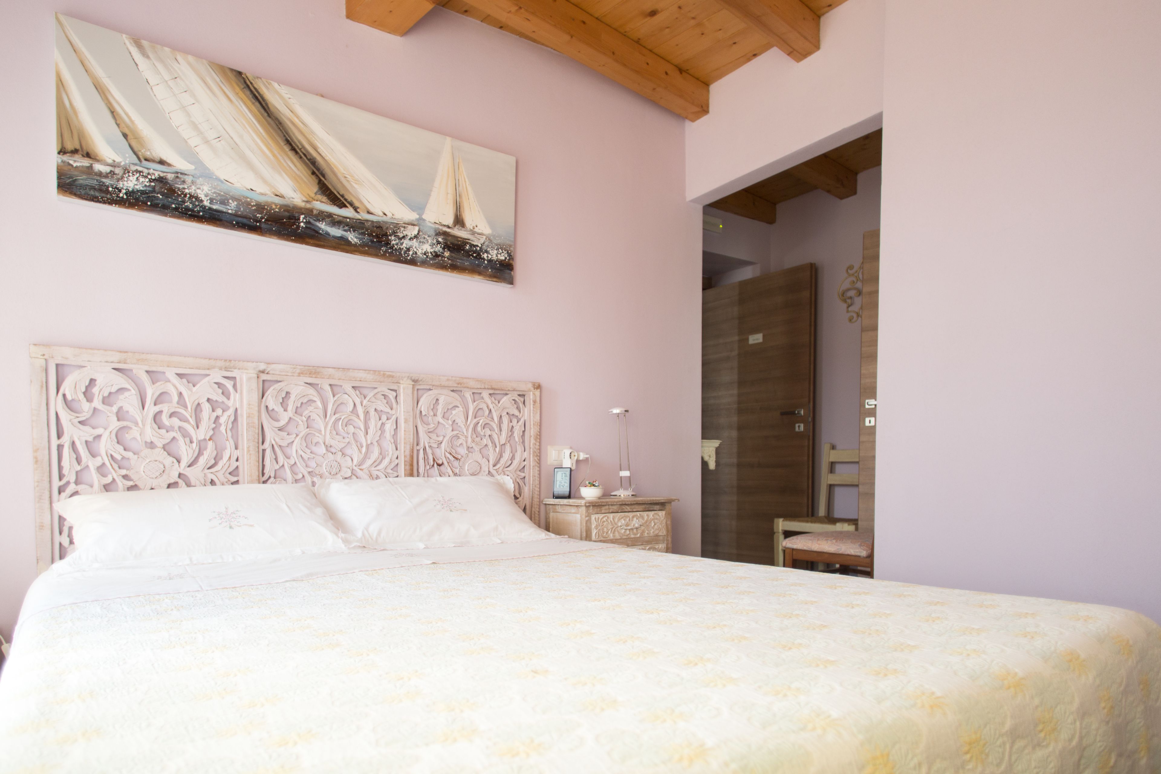 A bedroom with a large bed, a wooden ceiling, a painting of sailboats, a nightstand, and a chair.