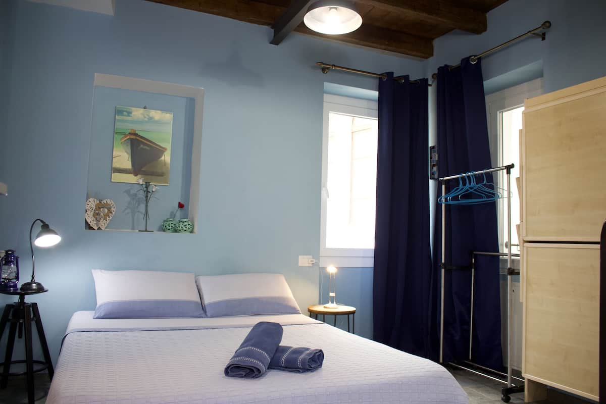 A neatly made bed with a rolled towel, a bedside lamp, a small table with a lamp, a window with curtains, and a wall-mounted drying rack.