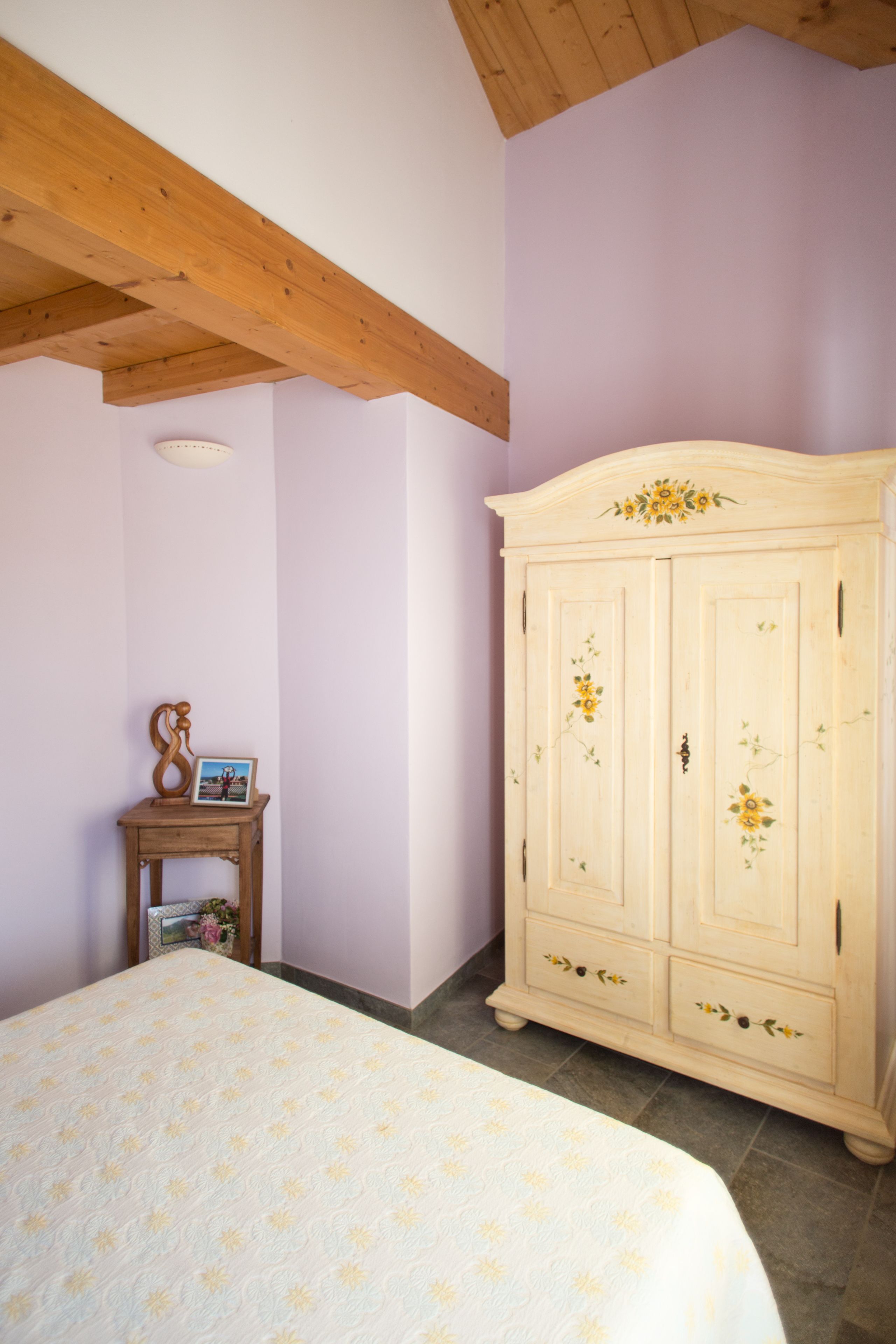 Een slaapkamer met een bed, een houten hoofdbord, een kledingkast en een kleine tafel met een fotolijst en vaas.