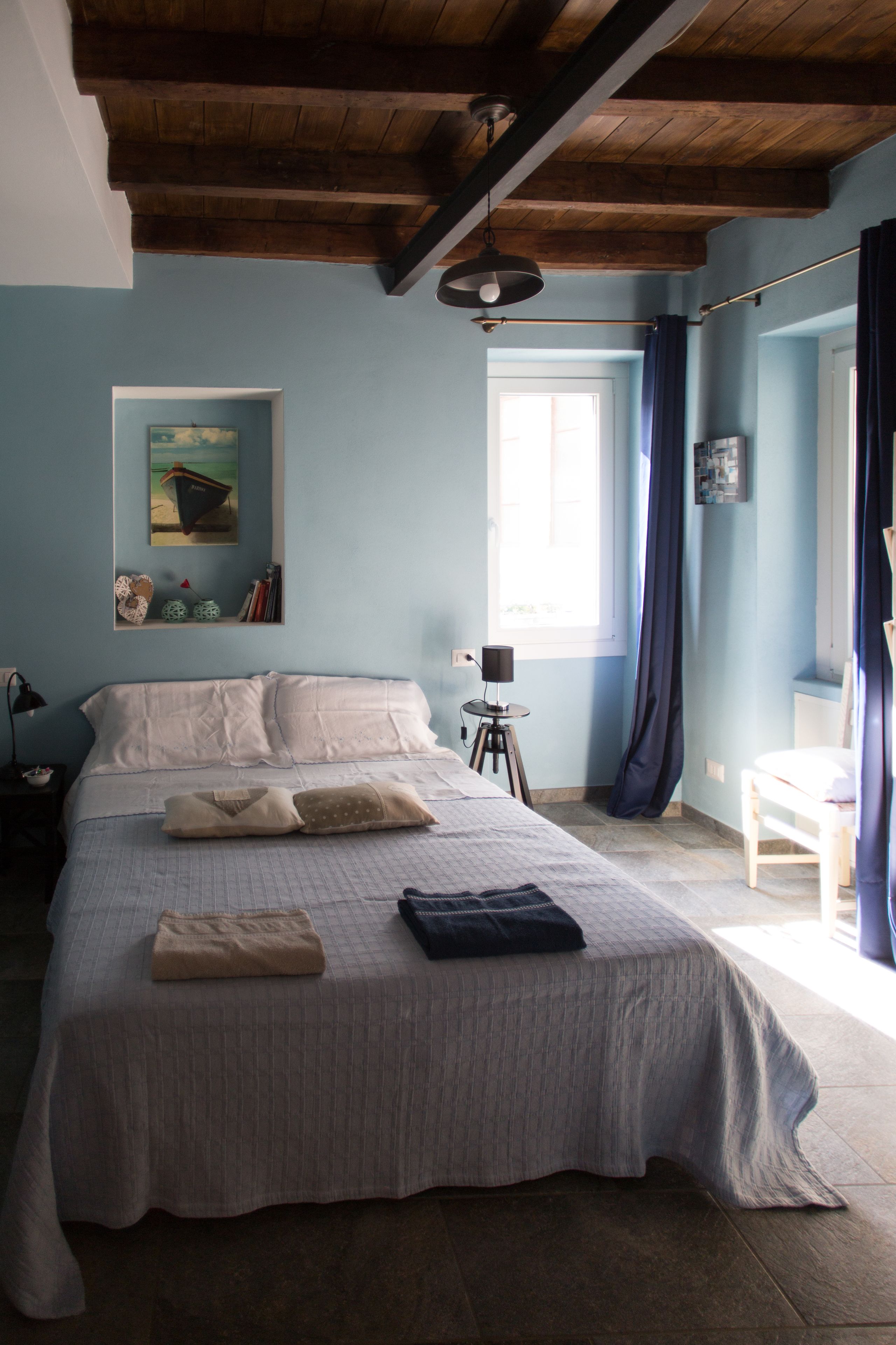A bedroom with a large bed, a small table, a window with curtains, and a ceiling with exposed beams.