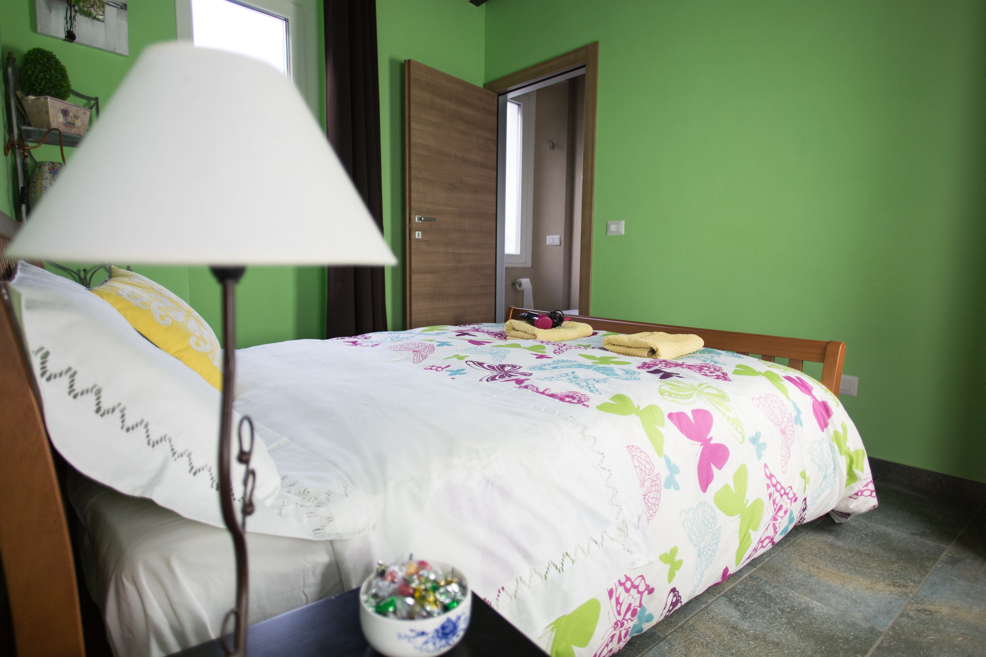 Ein Schlafzimmer mit einem Bett, einer Nachttischlampe, einem kleinen Tisch mit einer Schale Süßigkeiten und einer Tür zu einem anderen Raum.