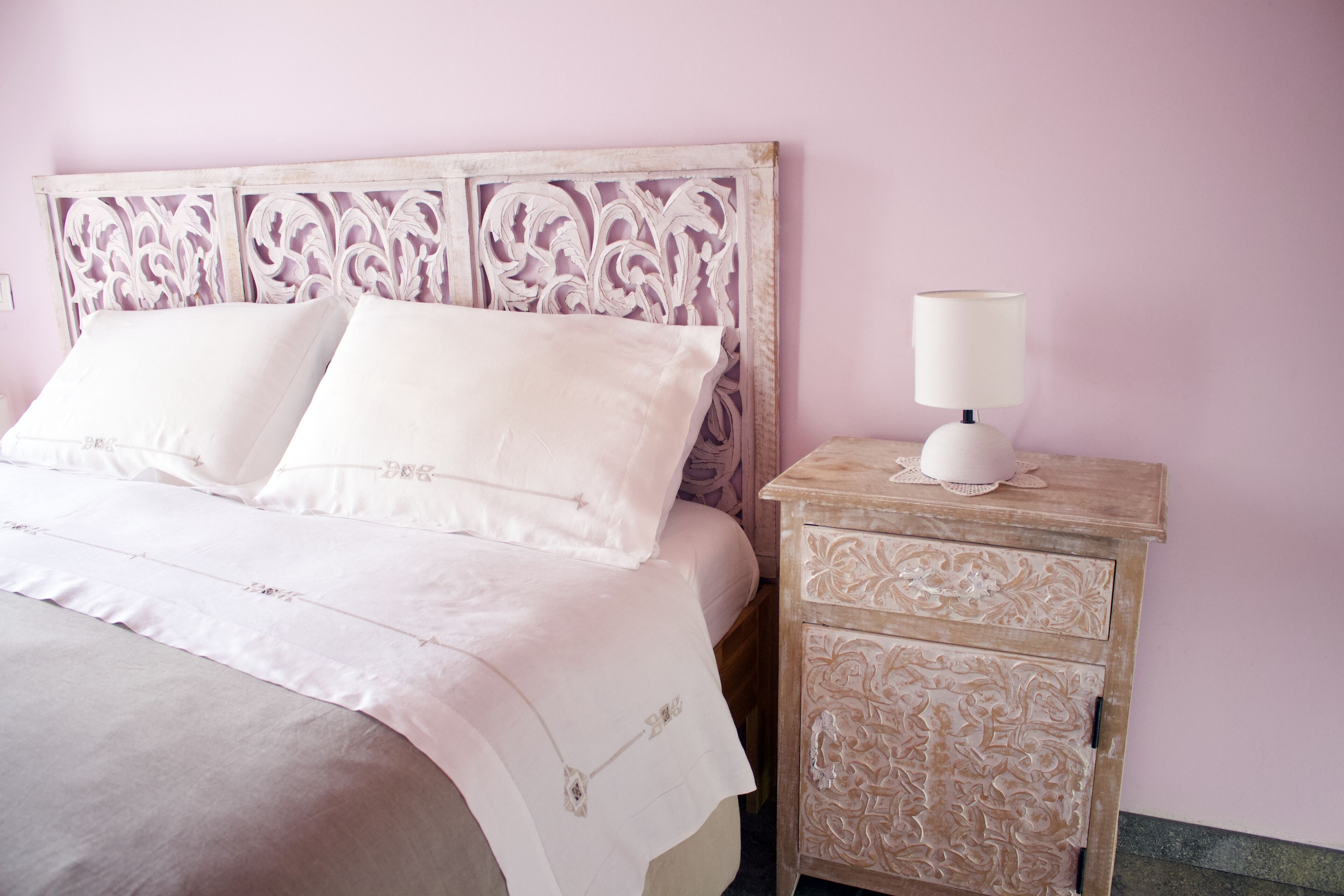 Una cama con ropa blanca y un cabecero decorativo, una mesita de noche con una lámpara y una pared con estampado floral.