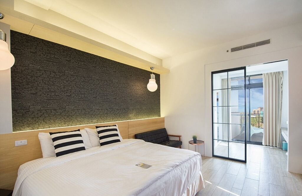 A modern hotel room with a large bed, a dark textured wall, a light fixture, and a sliding glass door to a balcony.