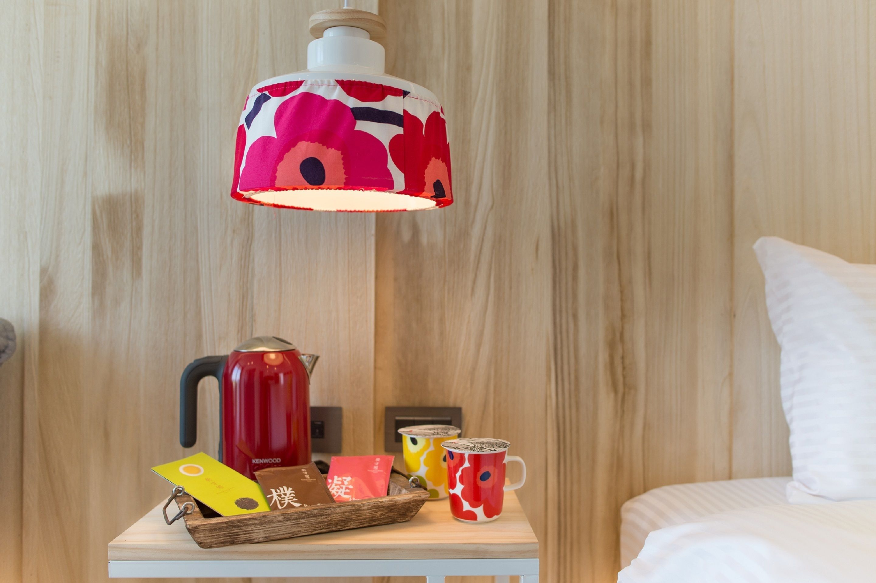 A wooden wall, a bed with white bedding, a nightstand with a red kettle, a colorful lamp, and a tray with cards and a mug.