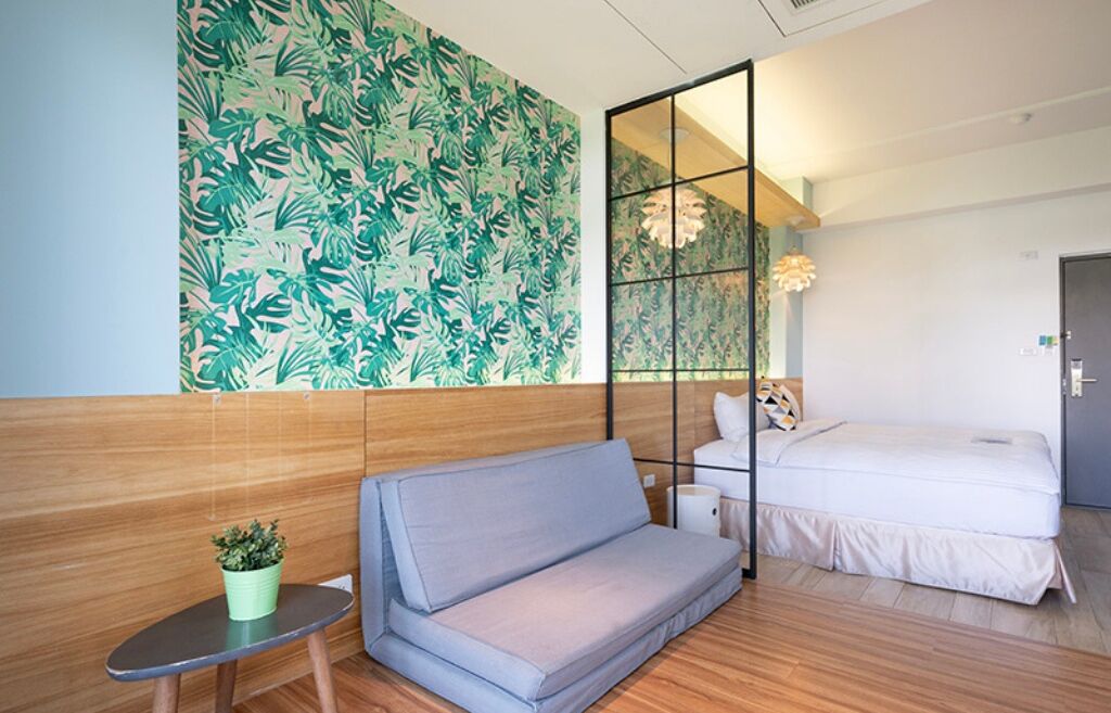A hotel room with a wooden floor, a grey sofa, a bed with white bedding, a small table with a potted plant, and a large window with a view of the outdoors.