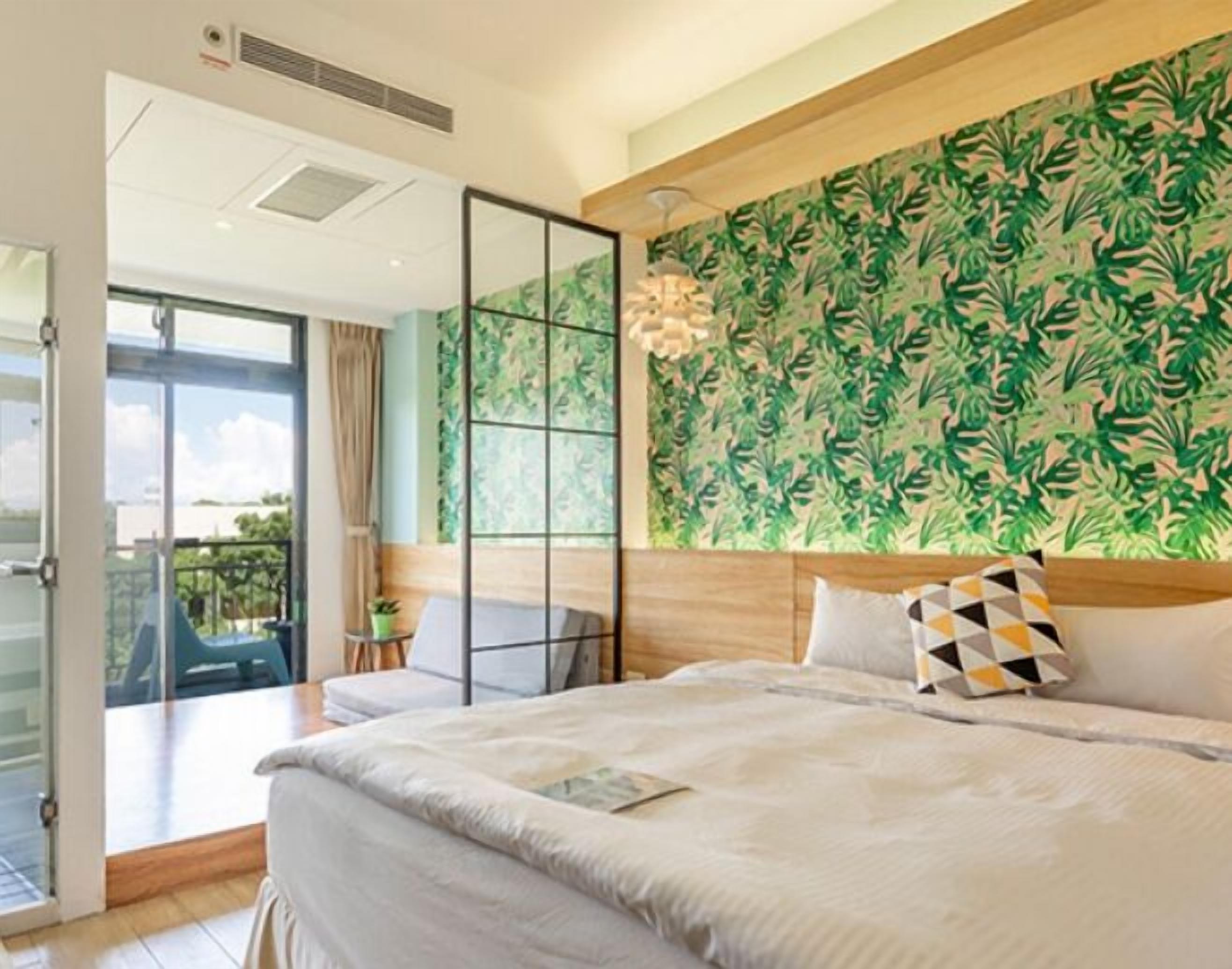 A bedroom with a large bed, a view through a sliding glass door, a green leafy wallpaper, and a ceiling fan.