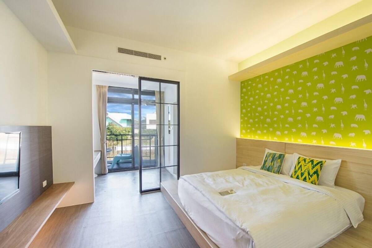A hotel room with a bed, a desk, a sliding glass door leading to an outdoor area, and a wall with a green animal pattern.