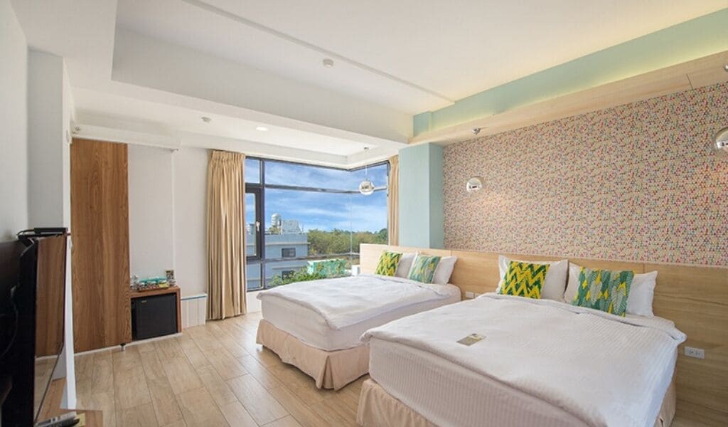 Habitación de hotel con dos camas, un armario de madera, una mesita pequeña y un ventanal con vistas a la ciudad.
