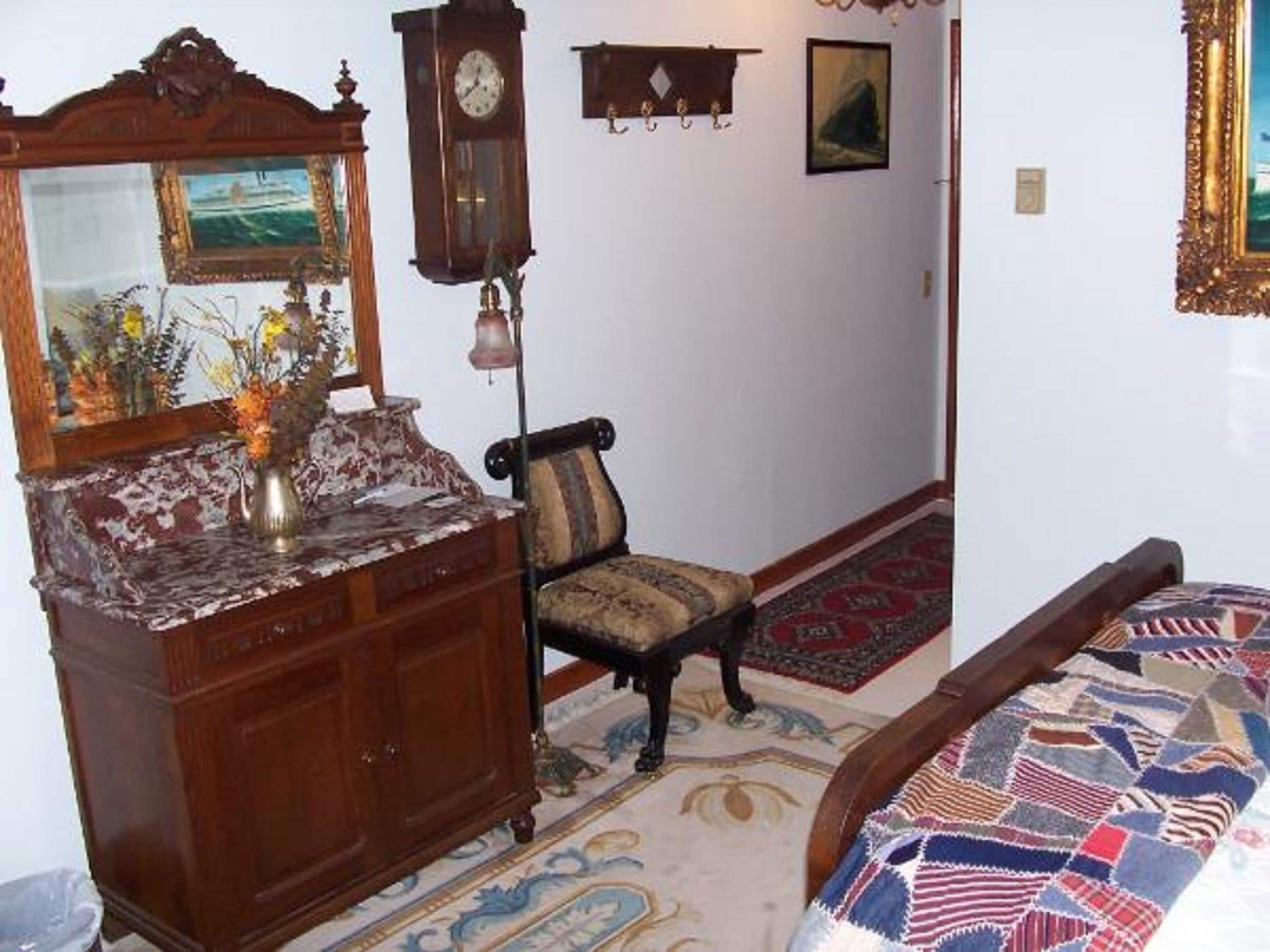 Een kamer met een houten dressoir, een stoel, een bed met een decoratief beddegoed, een klok en een schilderij aan de muur.