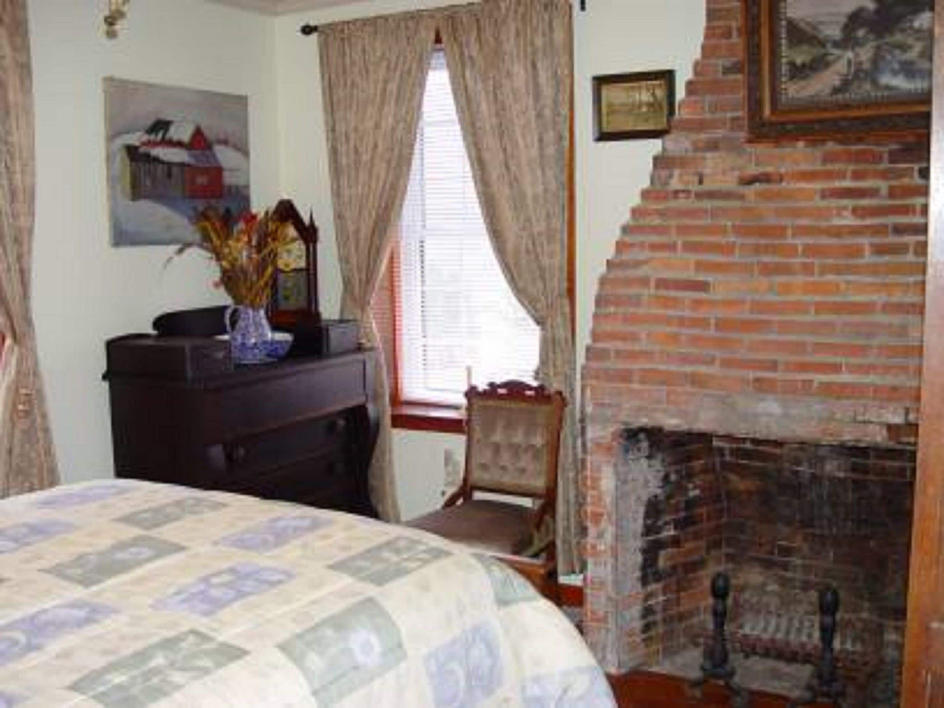 Een kamer met een open haard van baksteen, een bed met een sprei, een dressoir en een raam met gordijnen.