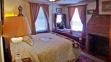 A bedroom with a bed, a nightstand, a lamp, a mirror, a fireplace, and a window with curtains.