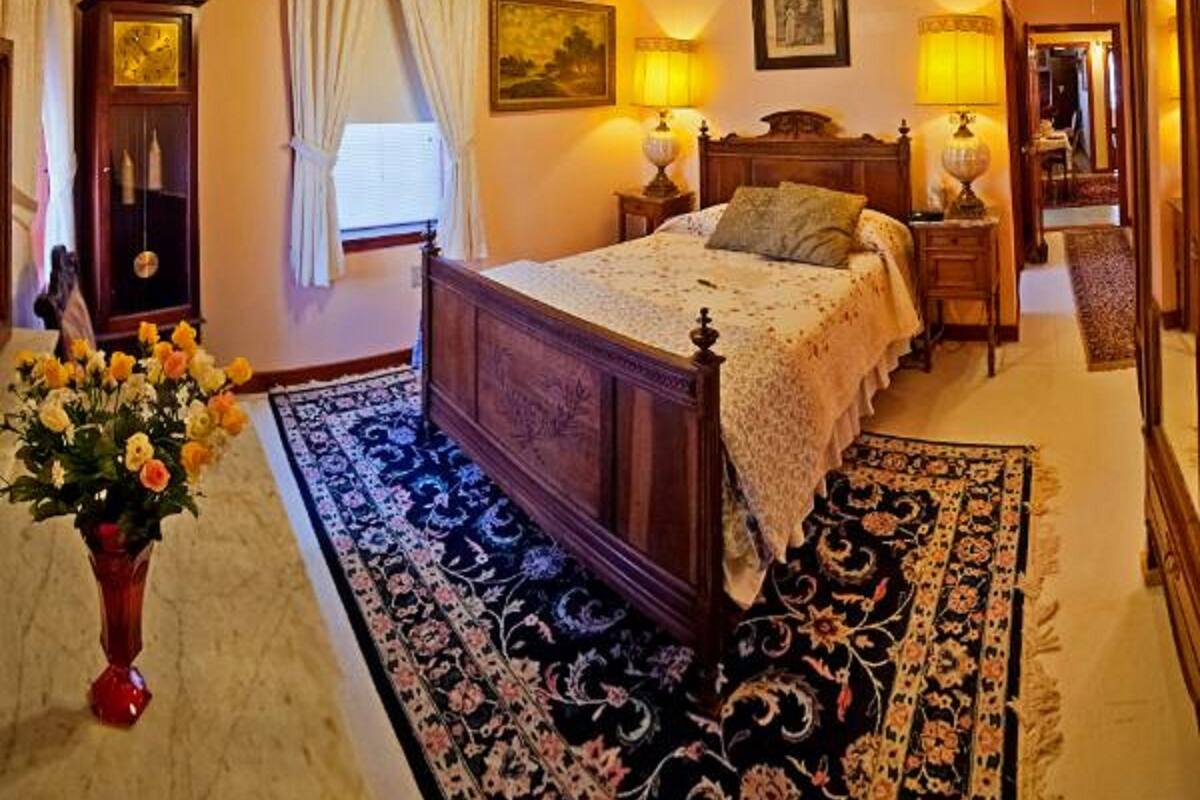 A bedroom with a large bed, a wooden wardrobe, a nightstand, a lamp, a painting on the wall, and a mirror.