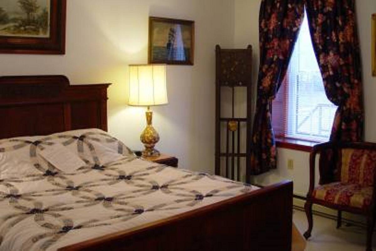 A bedroom with a bed, a nightstand with a lamp, a chair, and two framed pictures on the wall.