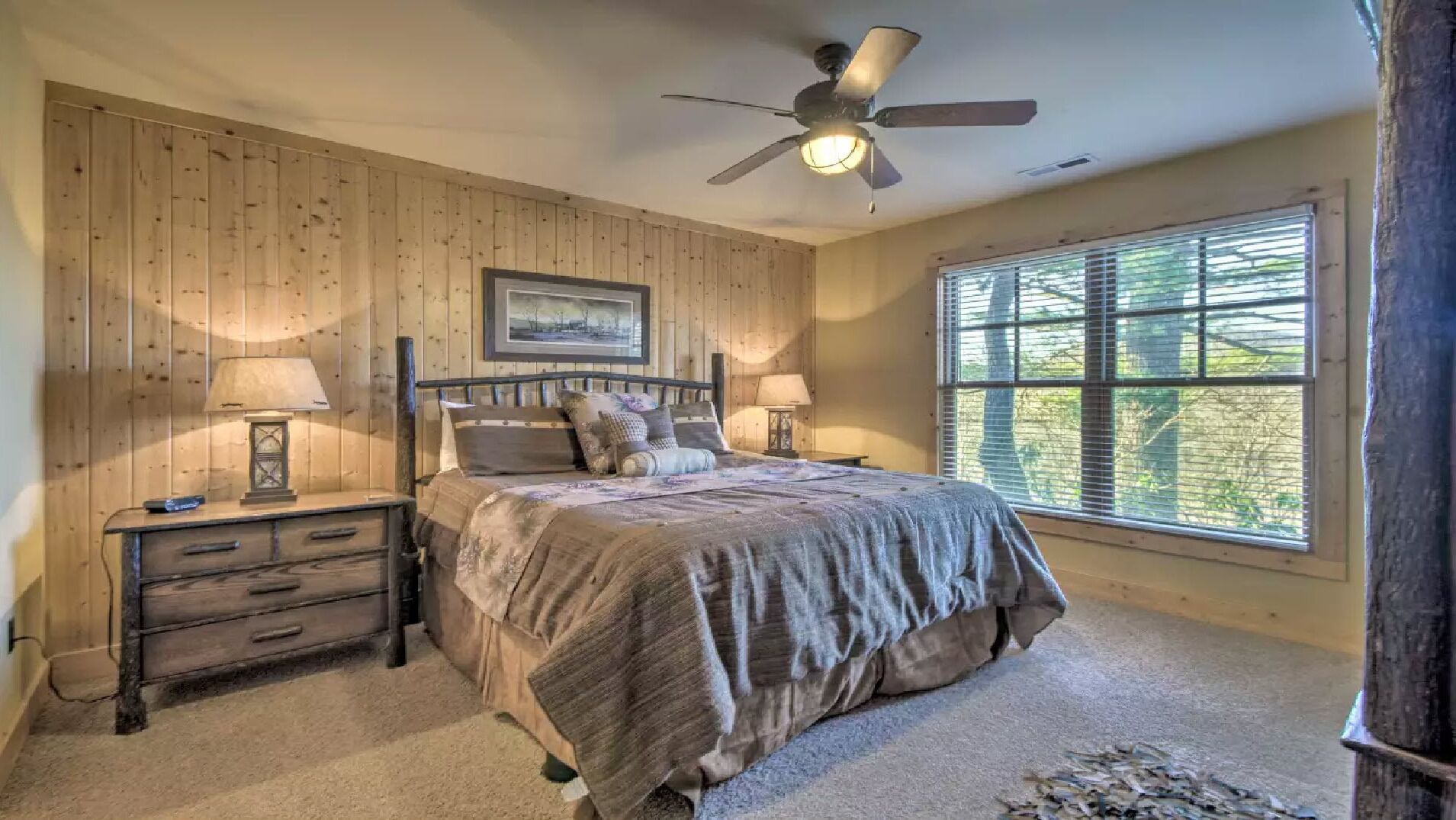 Une chambre à coucher comprenant un lit, deux tables de chevet, un ventilateur de plafond, une fenêtre avec des stores et un tableau encadré accroché au mur.