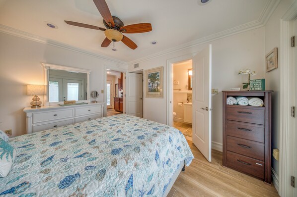 Une chambre à coucher avec un lit, une commode, un ventilateur de plafond et une salle de bain visible à travers une porte ouverte.