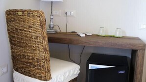 Una habitación con una silla de mimbre, un escritorio con lámpara, un refrigerador negro pequeño y un televisor montado.