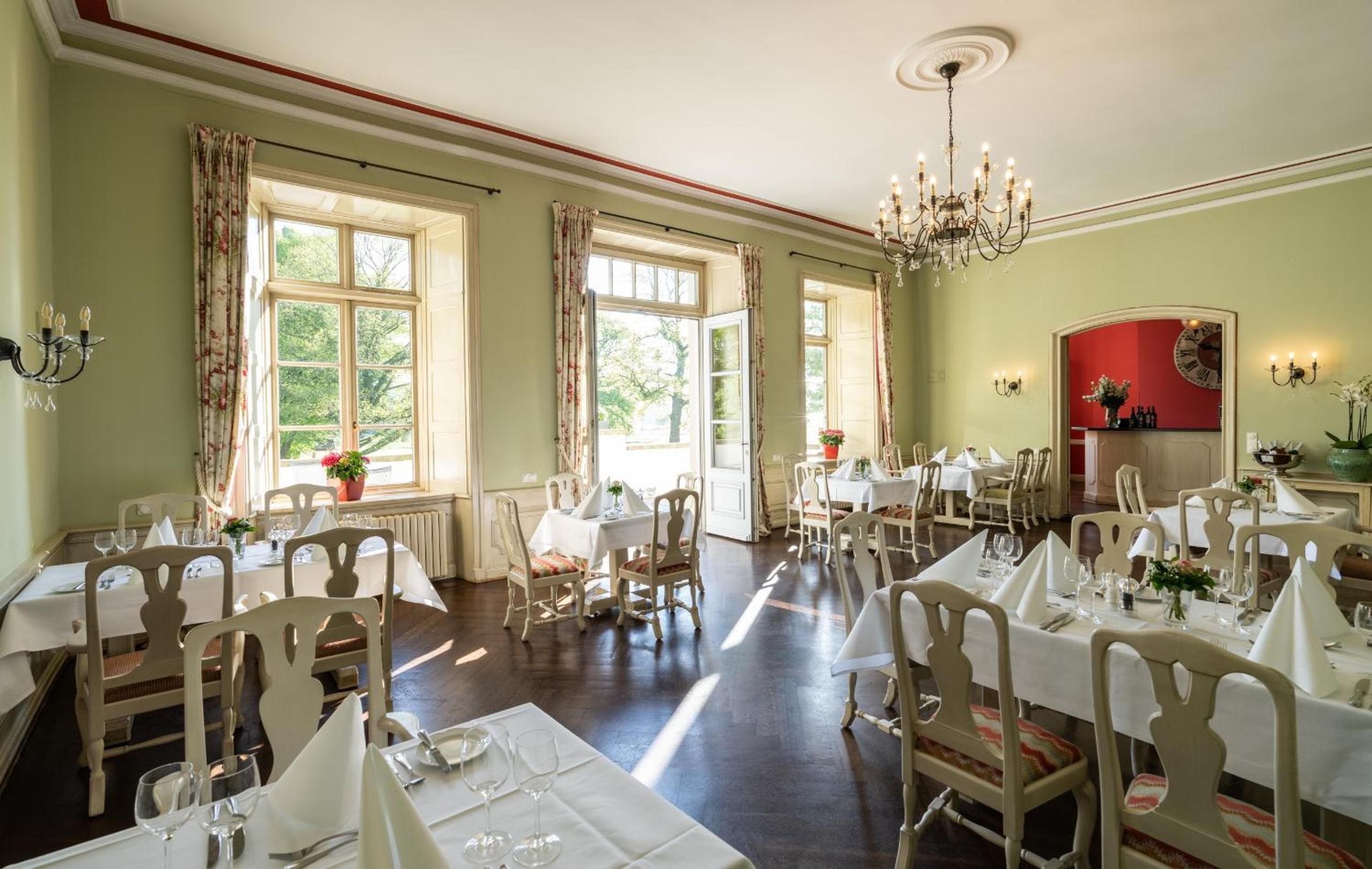 Un comedor con varias mesas dispuestas para el servicio, ventanas amplias con cortinas y un candelabro.