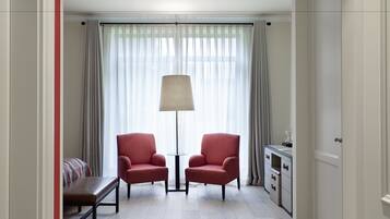 A hotel room with two red armchairs, a bench, a desk, and a lamp.