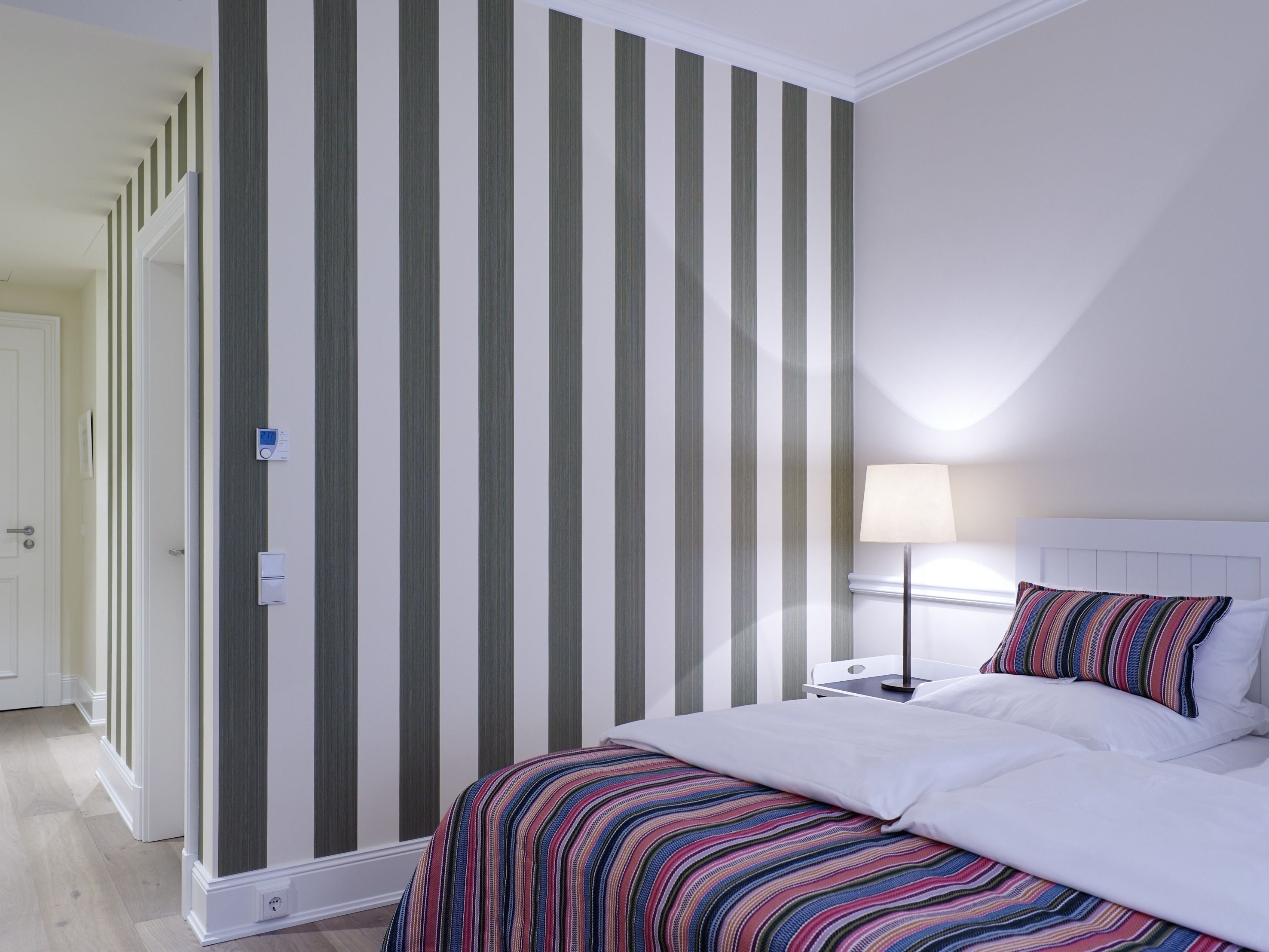 Un dormitorio con una pared a rayas, una cama con un edredón colorido a rayas, una mesita de noche con una lámpara y una puerta a otra habitación.