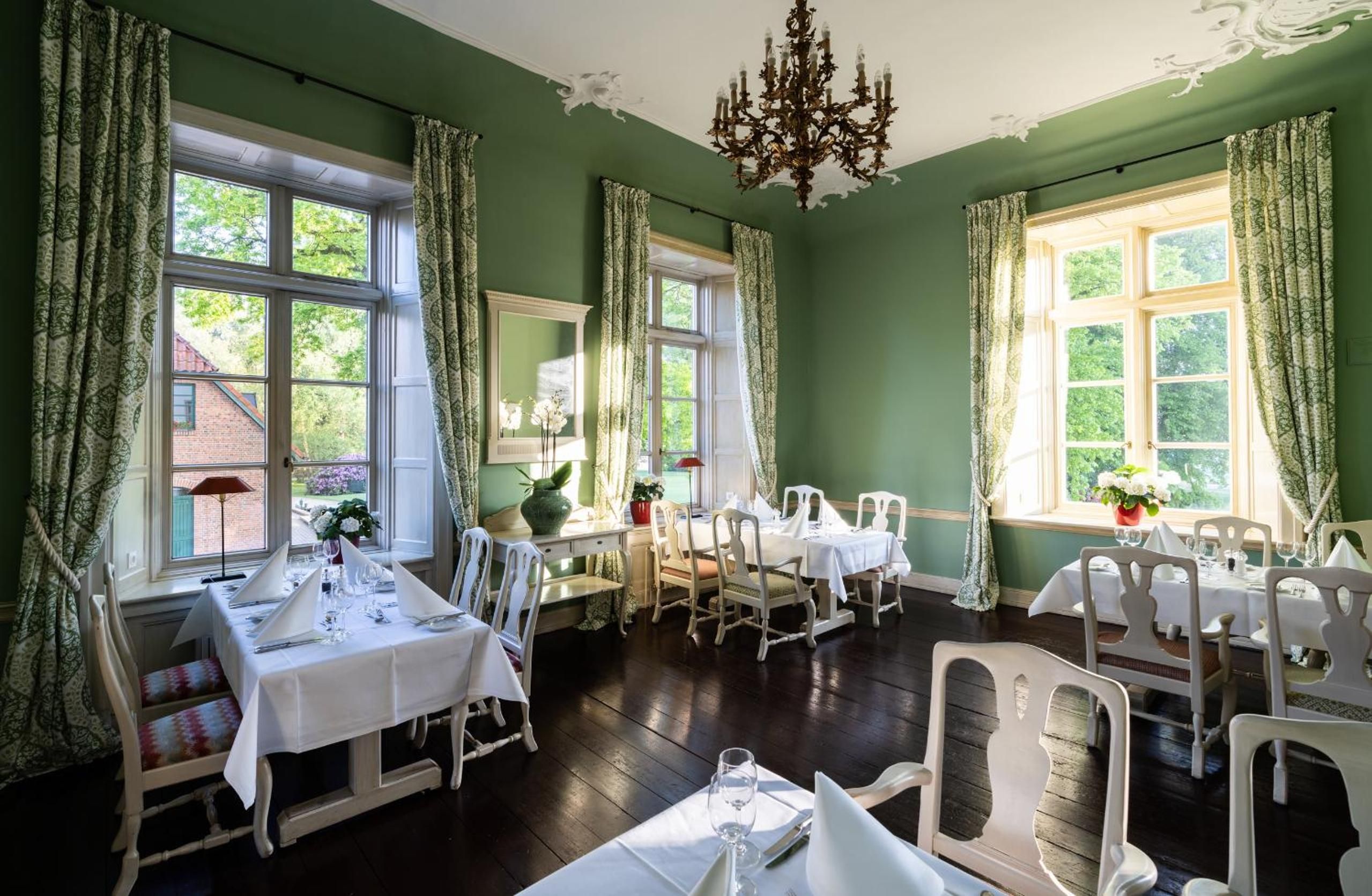Comedor con varias mesas dispuestas para una comida, ventanas amplias con cortinas y un candelabro.