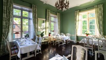 Comedor con varias mesas dispuestas para una comida, ventanas amplias con cortinas y un candelabro.