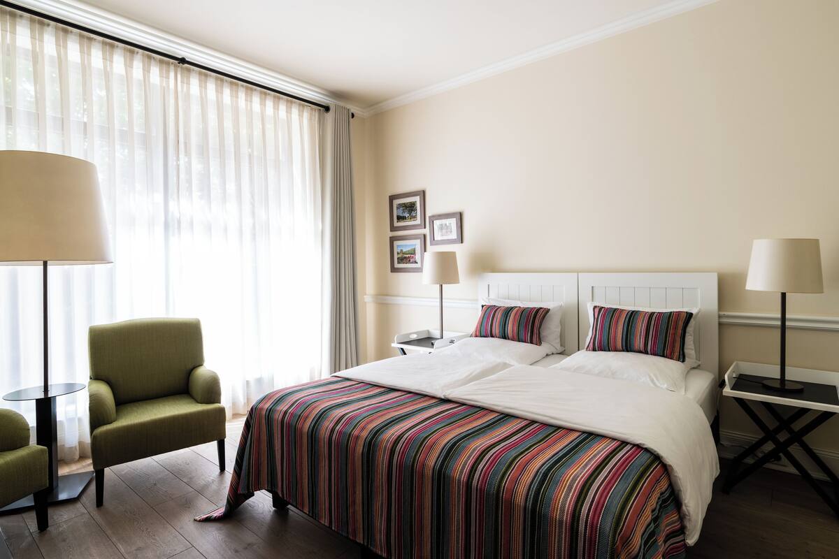 Una habitación de hotel con una cama grande, un sillón verde, dos mesitas de noche con lámparas y un ventanal con cortinas translúcidas.