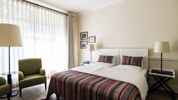 Una habitación de hotel con una cama grande, un sillón verde, dos mesitas de noche con lámparas y un ventanal con cortinas translúcidas.