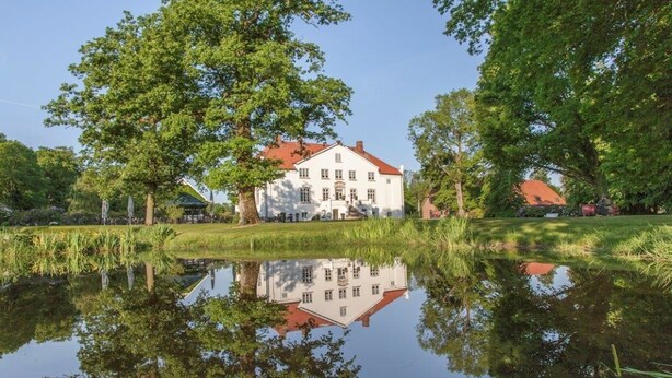 Een groot wit gebouw met een rood dak, omgeven door bomen en een rustig vijver waarin het gebouw wordt weerspiegeld.