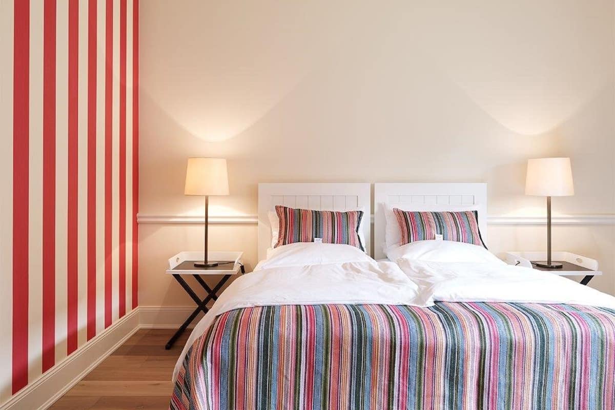 Habitación de hotel con dos camas, ropa de cama a rayas, pared con rayas rojas y blancas, y lámparas de noche.