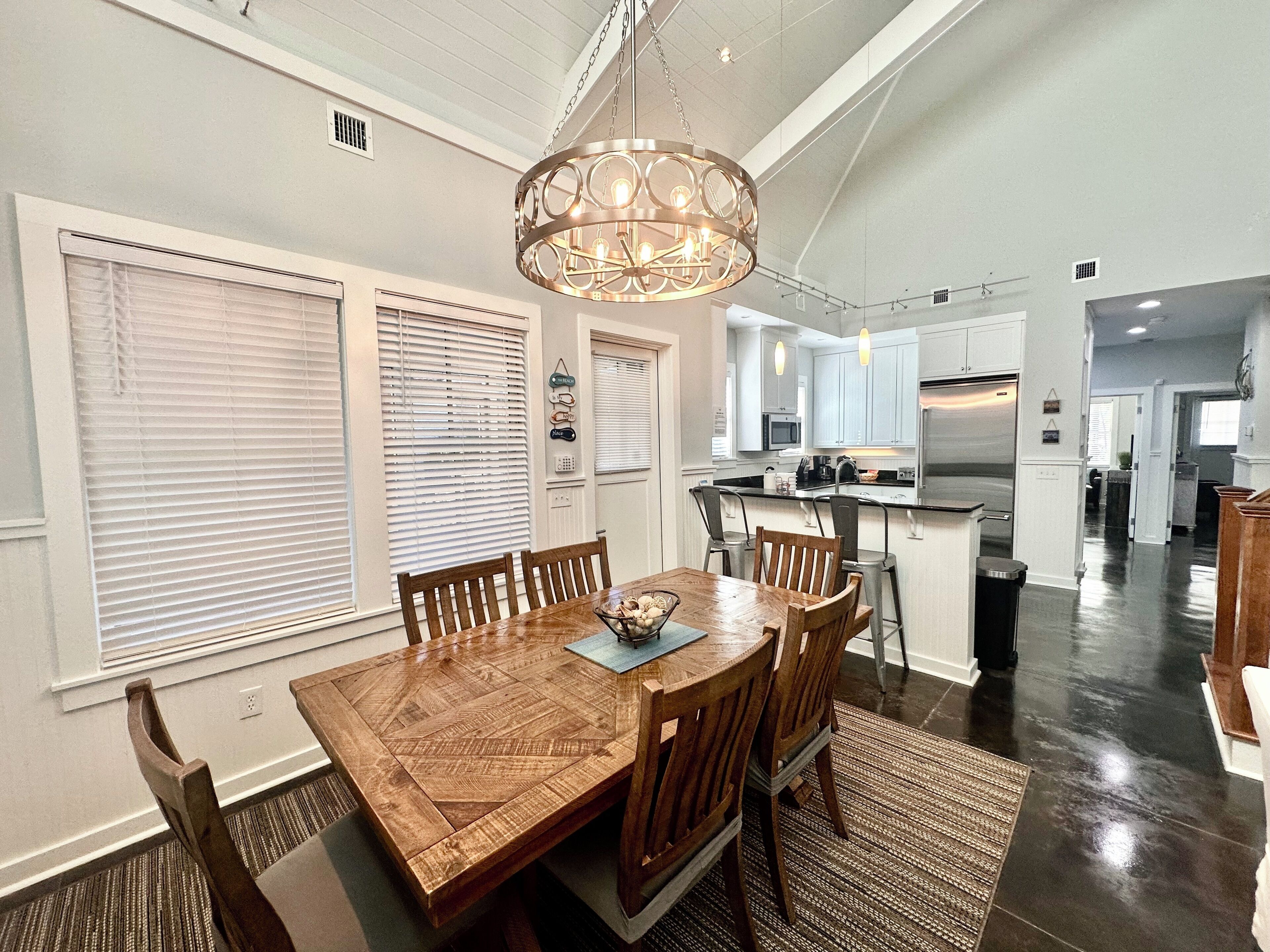 A dining area with a wooden table and chairs, a chandelier, and a kitchen in the background.