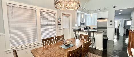 A dining area with a wooden table and chairs, a chandelier, and a kitchen in the background.