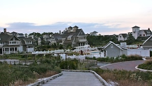 A wooden boardwalk leading to a beach area with houses and a white picket fence.