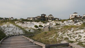 A wooden boardwalk leading to a row of beach houses with sandy dunes and vegetation.