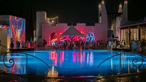 A pool area with a stage and audience, surrounded by buildings with illuminated facades.