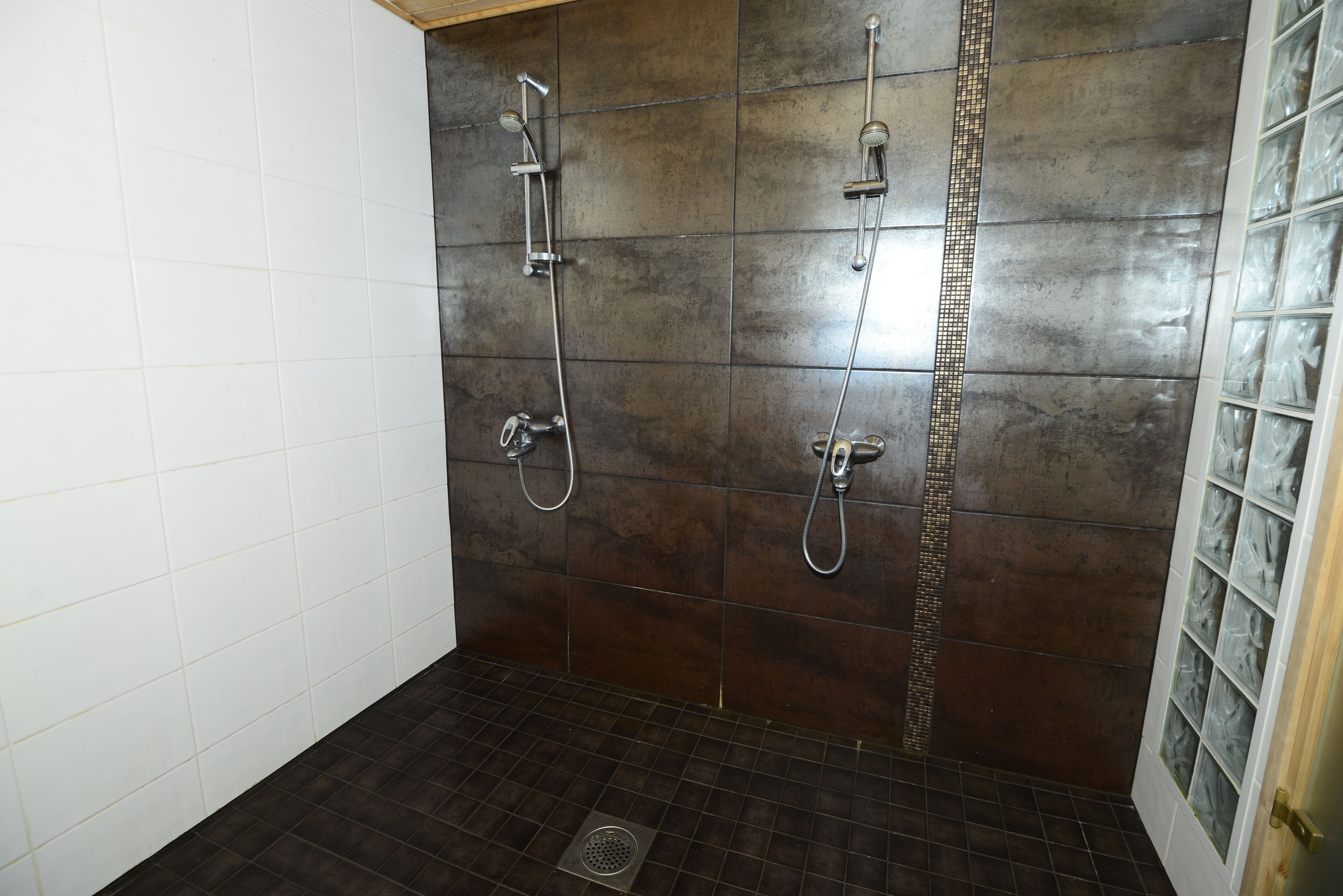 Área de chuveiro com azulejos escuros, parede revestida de azulejos brancos e porta de vidro com painéis texturizados.